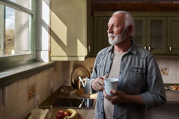 Senior man day dreaming while drinking coffee and looking through window at home.